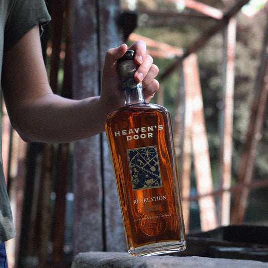 Person holding a bottle of Heaven's Door Revelation whiskey outdoors.