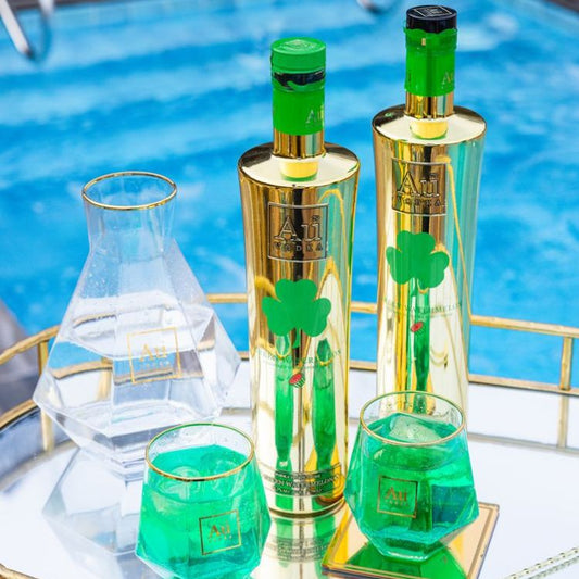 Two green bottles with gold accents and glasses filled with a green liquid on a reflective surface.