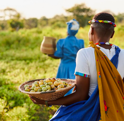 Two women in traditional attire holding a basket of fruits and a pot in an outdoor setting.