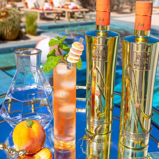 Two bottles of Aji liqueur with a cocktail and peach by a poolside.