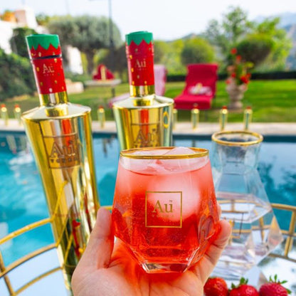 Hand holding a red cocktail in a gold-rimmed glass with 'Au' branding, next to two gold bottles in an outdoor setting.