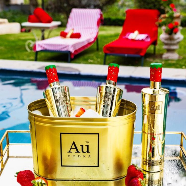 Gold Au Vodka bottle and shaker set by a poolside with lounge chairs and flowers.