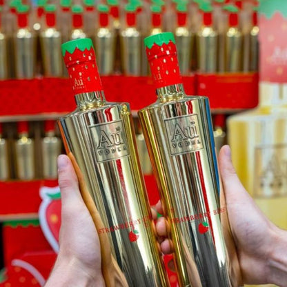 Two gold bottles with strawberry designs held by a person against a blurred background of similar bottles.