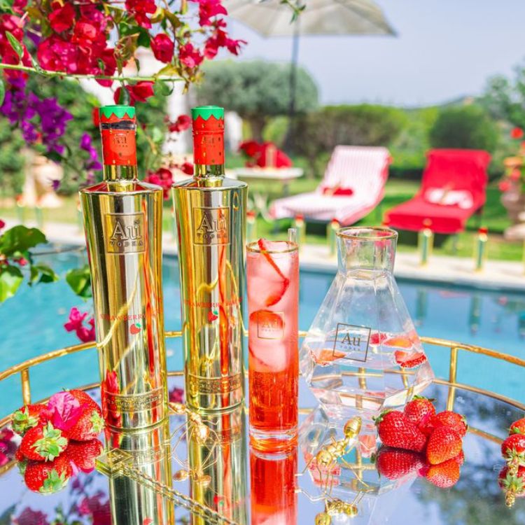 Two gold bottles with red labels on a table by a poolside with flowers and drinks.