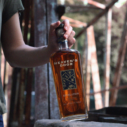 Person holding a bottle of Heaven's Door Revelation whiskey outdoors.
