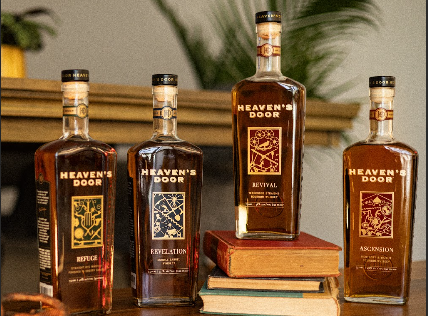 Four bottles of Heaven's Door whiskey on a wooden surface with books in the background.