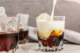 Glass of iced coffee with cream being poured into it, with another glass of iced coffee in the background.