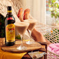 Two glasses of a creamy cocktail with a bottle of Amaretto on a wooden tray, with a person lounging in the background.