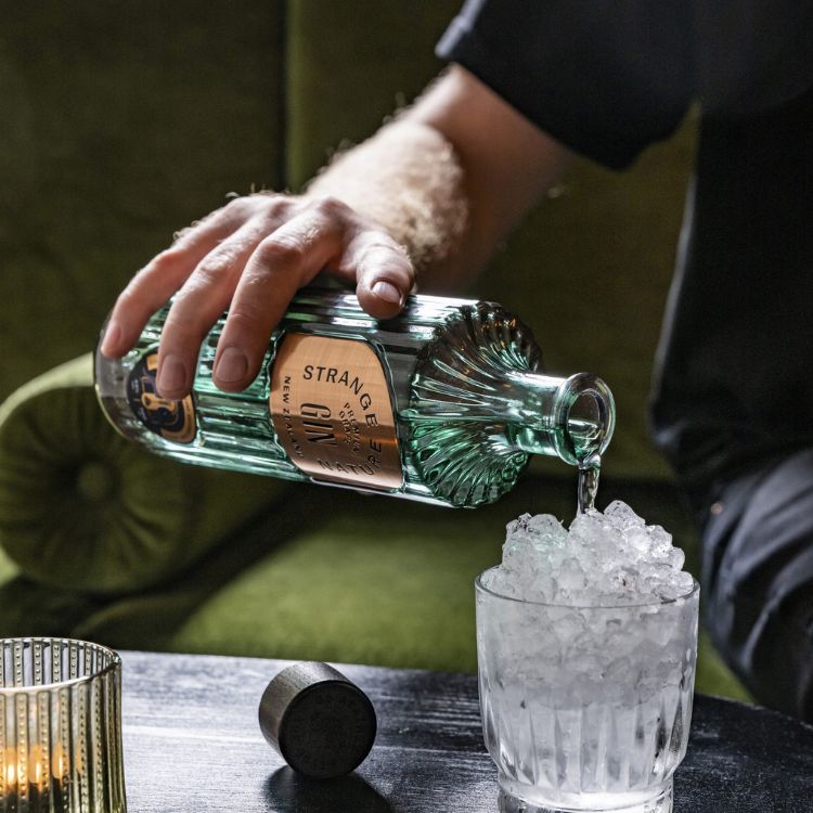 Person pouring Strange Gin into a glass with ice on a wooden table.