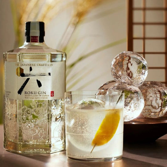 Rokugin Japanese craft gin bottle with a glass of gin and tonic garnished with a lemon peel, against a decorative background.